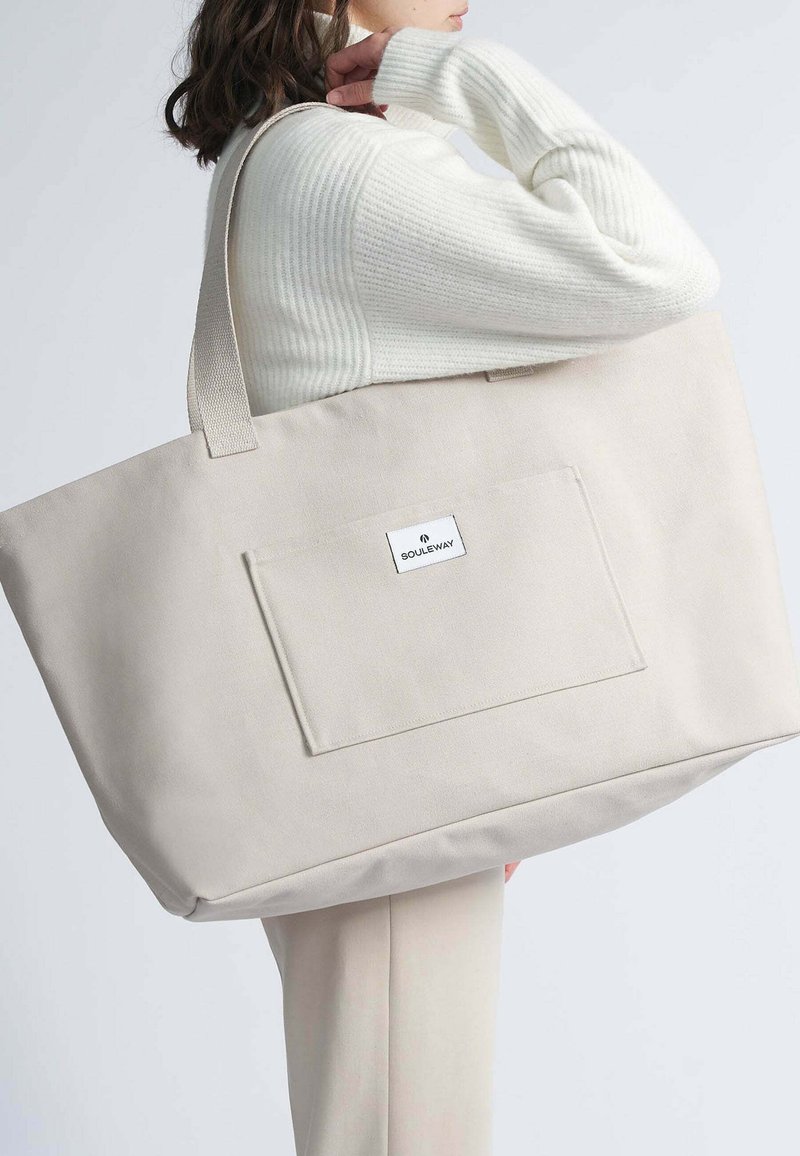 Bolsa tote de lienzo beige con dos asas resistentes y un bolsillo frontal. Presenta una etiqueta de marca en el bolsillo y una textura suave y duradera.