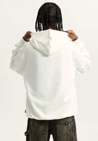 Hoodie bianco oversize con cappuccio regolabile, polsini a coste e texture liscia; indossato con pantaloni scuri e texture che presentano tasche.