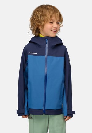 Smilende barn med krøllet hår iført en blå og marineblå Mammut jakke og lysegrønne bukser, kiggende mod højre.