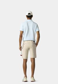Lichtblauwe polo, beige shorts, witte hoed met "POLO" logo, een bloem vasthoudend, en sportschoenen met kleurrijke accenten.
