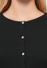 Top de punto negro con escote redondo, con tres botones decorativos plateados en forma de flor en la parte delantera. Textura suave, diseño casual.