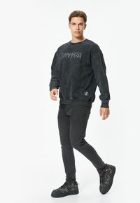 Sudadera negra con gráfico de "DARKNESS" en un estilo desgastado, combinada con jeans negros y zapatillas negras texturizadas, exhibida en un modelo.