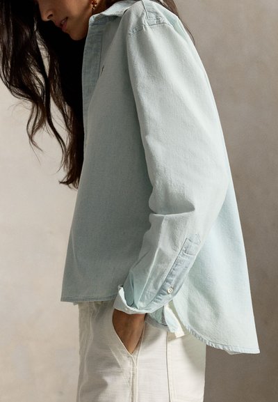 Kvinde med langt mørkt hår iført lyseblå oversized skjorte og hvide bukser, stående med den ene hånd i lommen mod en neutral baggrund.