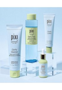 Fire Pixi hudpleieprodukter anordnet på klare pedestaler: Clarity Cleanser (hvit tube, mintfarger), Clarity Tonic (blå flaske, gul hette), Clarity Lotion (hvit tube) og Clarity Concentrate (grønn flaske, gylden pipette).