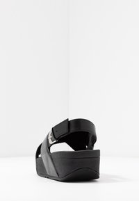 Svart kilklack sandal med justerbart ankelband och silver spänne, vinklad vy mot en slät vit bakgrund.