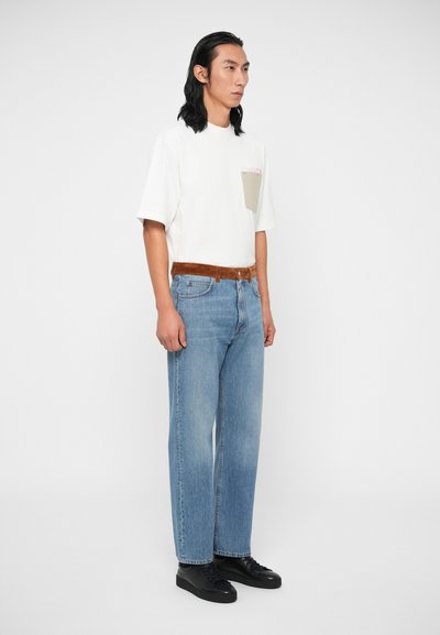 Fehér, rövid ujjú póló bézs mellzsebbel, világoskék, egyenes szárú farmernadrággal és fekete sportcipővel párosítva. Barna velúr öv emeli ki az összhatást.