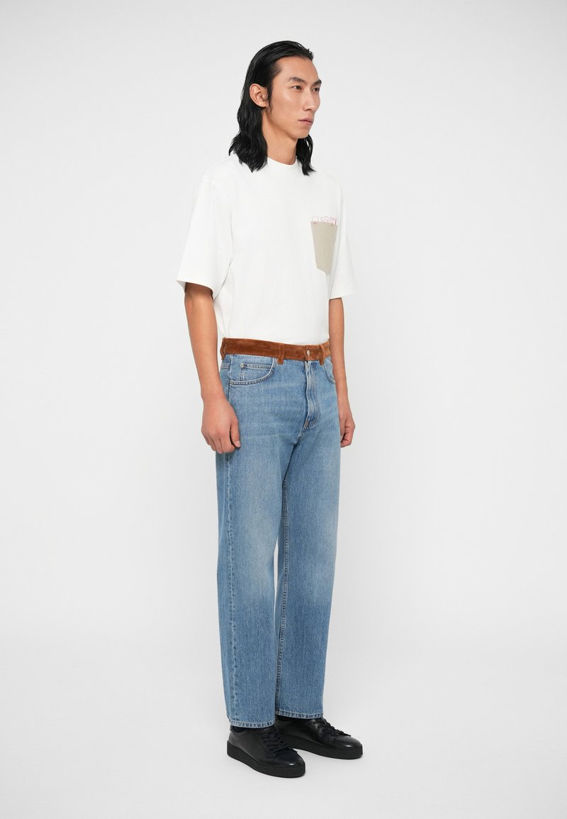 Weißes kurzärmliges T-Shirt mit einer beigen Brusttasche, kombiniert mit hellblauen Geradeausjeans und schwarzen Sneakers. Brauner Wildledergürtel setzt Akzente.
