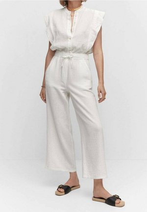 Frau trägt ärmellosen weißen, strukturreichen Overall mit Kordelzug in der Taille und schwarzen Sandalen zum Hineinschlüpfen, steht vor weißem Hintergrund.