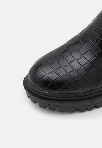 Bottine en cuir noir avec un motif tissé texturé, bout arrondi, semelle en caoutchouc avec crampons pour la traction, et finition cousue.