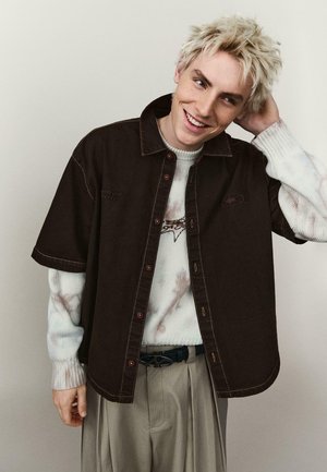 Mladý človek s krátkymi blond vlasmi sa usmieva, má na sebe tmavú krátkym rukávom košeľu zapnutú na gombíky cez svetlý dlhý rukáv sveter a béžové nohavice.