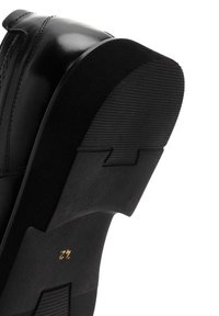 Chaussure en cuir noir avec une semelle en caoutchouc texturée, présentant une silhouette lisse et une subtile marque dorée visible sur le talon.