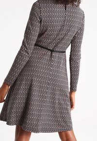 Femme portant une robe à manches longues et à motifs, longueur au genou, avec une taille cintrée et une jupe évasée, photographiée de dos.