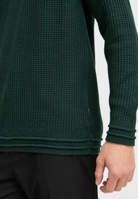 Pull en maille texturée vert foncé avec un motif en nid d'abeille, présentant des poignets et un ourlet côtelés, associé à un pantalon noir.