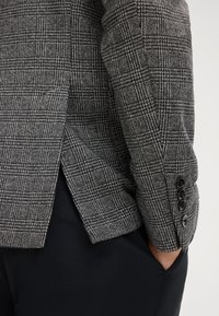 Grijze geruite blazer met een gestructureerde stof, notch lapel, zijsplit en drie-knopen manchetten, gedragen over zwarte broek.