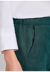 Teal leren broek met een plooidetail en een elastische tailleband, gecombineerd met een witte gestructureerde lang- mouw shirt. Zichtbare zakopening.