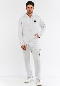 Haut à capuche zippé gris clair avec un logo noir rond sur la poitrine, associé à un pantalon de survêtement assorti avec une impression graphique noire.