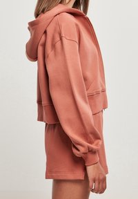 Felpa corta con cappuccio in rosa polvere, realizzata in tessuto morbido. Presenta una zip frontale, cappuccio e polsini elasticizzati. Abbinata a pantaloncini coordinati.