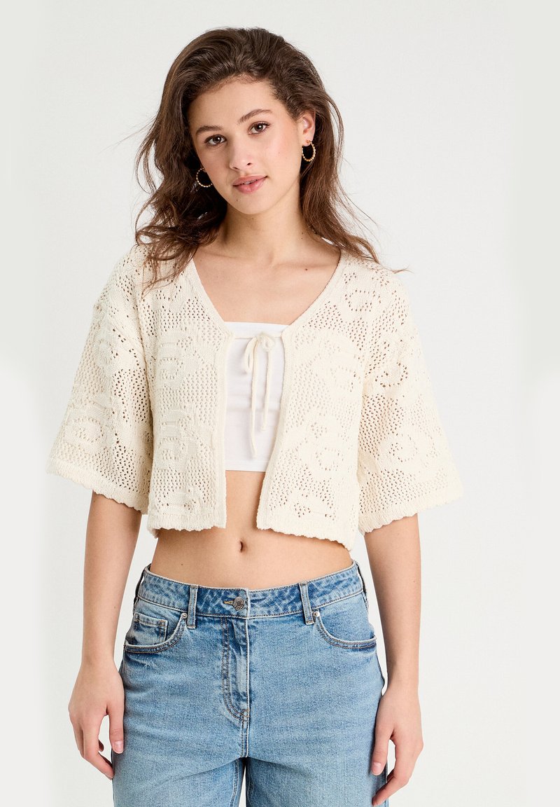 Un cardigan en crochet crème avec un col en V, des manches courtes et des bords festonnés, associé à un crop top blanc et un jean bleu clair.
