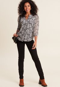 Schwarz-weiß gemusterte Bluse mit dreiviertellangen Spitzensäumen, kombiniert mit schwarzen, eng anliegenden Hosen und rostfarbenen Ankle-Boots.