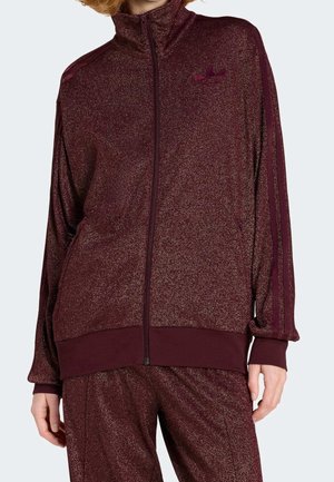 Vrouw draagt een glinsterend donkerrood zip-up sportjack met bijpassende broek, met gestreepte mouwen en een logo op de borst.