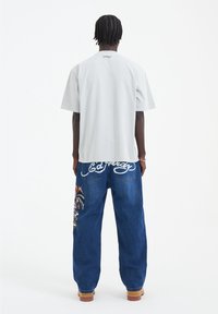 Jasnoszary oversize t-shirt z krótkimi rękawami, zestawiony z niebieskimi dżinsami z kolorową grafiką i białym haftowanym nazwiskiem marki na plecach.