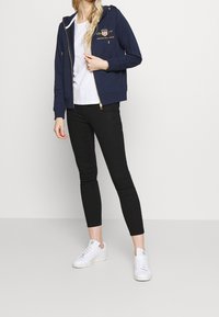 Námořnická mikina na zip se zlatými akcenty a logem, doplněná o bílý tričko, černé skinny džíny a bílé tenisky. Hladká tkanina a přiléhavý střih.