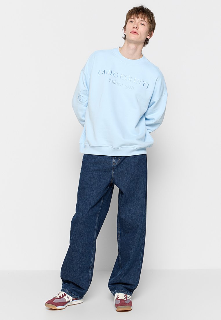 Lichtblauwe sweatshirt met reliëflogo, gecombineerd met loszittende donkere denim jeans en multicolor sneakers. Casual stedelijke stijl.