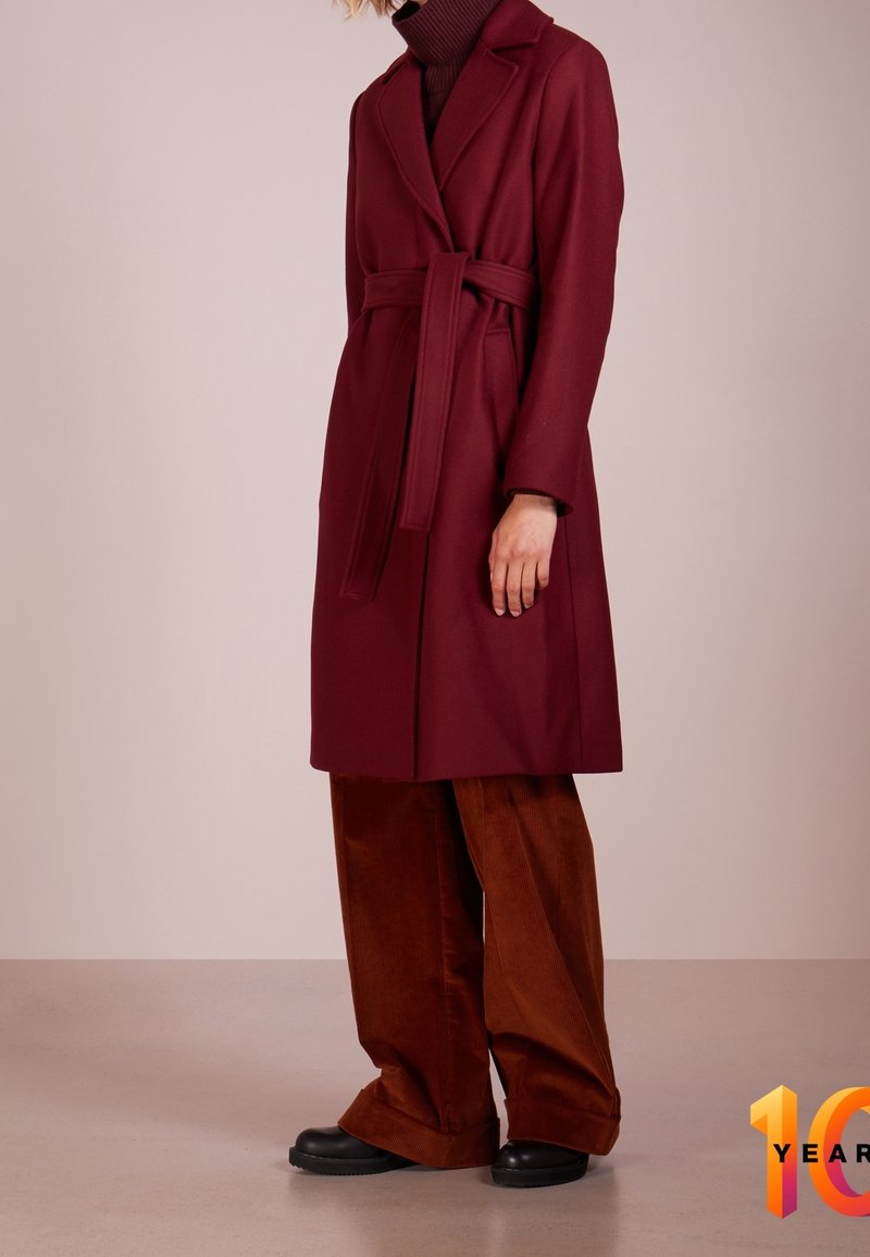 Abrigo de lana burdeos con cintura ceñida, solapa recortada y mangas largas, combinado con pantalones anchos de pana marrón y zapatos negros.