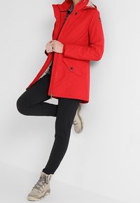 Parka roja con capucha, que cuenta con una cremallera de doble sentido, bolsillos laterales y un forro interior beige en contraste. Combinada con leggins negros y botas color tostado.