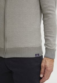 Grauer gestrickter Pullover mit gerippten Bündchen und Saum, der ein strukturiertes Muster und ein kleines Stoffetikett an der unteren Seite aufweist.