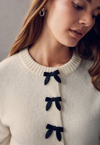 Cremefarbener Strickcardigan mit geripptem Kragen und drei schwarzen Schleifenakzenten. Weiche Textur, Knopfleiste vorne.
