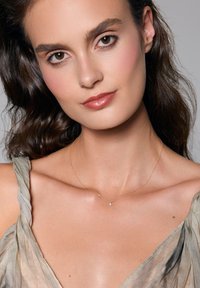 Złoty łańcuszek z małym zawieszką w kształcie gwiazdki. Modelka ma na sobie miękką, luźną bluzkę w jasnym odcieniu. Gładka skóra i naturalny makijaż.