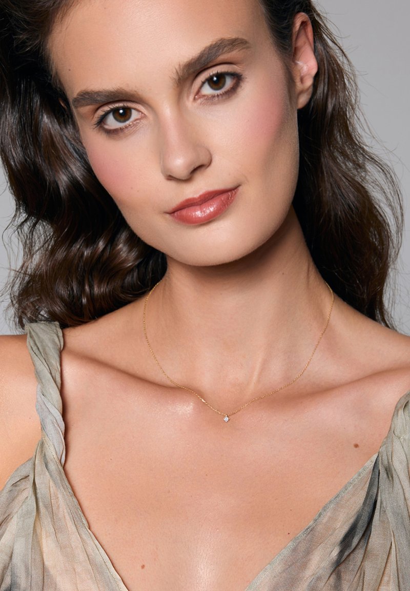 Złoty łańcuszek z małym zawieszką w kształcie gwiazdki. Modelka ma na sobie miękką, luźną bluzkę w jasnym odcieniu. Gładka skóra i naturalny makijaż.