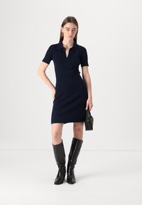 Abito a coste blu navy con maniche corte, colletto polo, vita aderente e orlo al ginocchio. Abbinato a stivali neri al ginocchio e una borsa piccola.