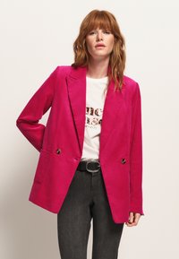 Blazer in corduroy fucsia con design doppiopetto, revers a punta, due tasche frontali e dettagli con bottoni dorati. Indossato sopra una t-shirt con grafica.