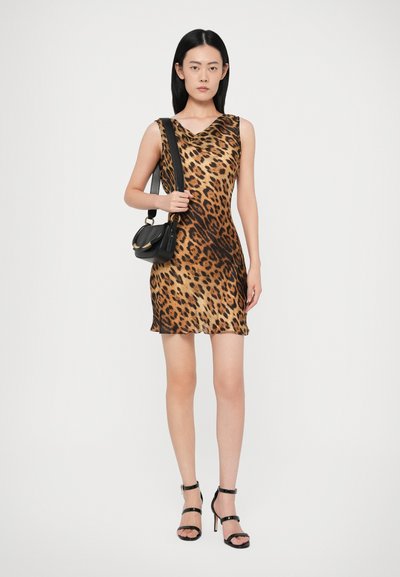 Mulher vestindo um vestido sem mangas com estampa de leopardo e sandálias de salto alto pretas, segurando uma bolsa preta de ombro contra um fundo simples.