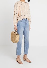 Blusa pescoza con ricamo a forma di farfalla, jeans azzurri, sandali beige con tacco e borsa di paglia intrecciata. Vestibilità comoda, tessuto morbido, stile casual.