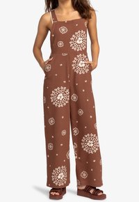 Bruine jumpsuit met een patroon van witte bloemen, wijde benen, verstelbare schouderbanden en zijzakken. Gecombineerd met donkere platform sandalen.