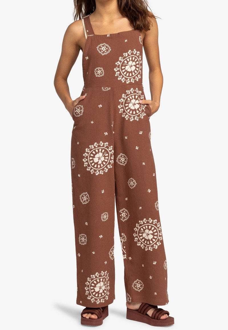 Bruine jumpsuit met een patroon van witte bloemen, wijde benen, verstelbare schouderbanden en zijzakken. Gecombineerd met donkere platform sandalen.