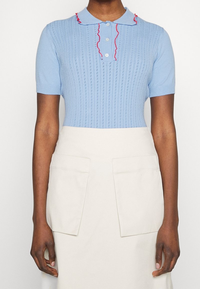 Light blue short-sleeve knitted polo with a textured pattern, white buttons, and red ruffled accents on the collar. Worn with a beige skirt.
