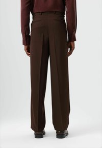 Bruine broek met wijde pijpen en een gladde textuur, voorzien van achterzakken en een getailleerde pasvorm. Gecombineerd met een bordeauxrode shirt met lange mouwen.