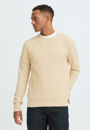 Maglione beige chiaro lavorato a maglia con motivi verticali strutturati, scollo rotondo e maniche lunghe. Abbinato a jeans scuri.