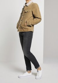 Person som bär en beige manchestersak med fårskinnskrage, svarta skinny jeans och vita sneakers, står med händerna i fickorna.