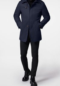 Manteau imperméable bleu marine avec des boutons en accent, des épaulettes et des poches avant. Associé à un pantalon noir slim et des bottes noires épaisses.