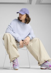 Sweat-shirt bleu clair, pantalon large beige et casquette violette. Les baskets présentent des accents gris, violets et roses avec un design texturé.