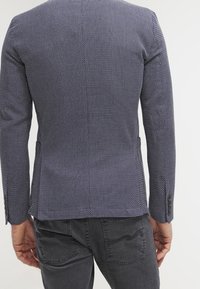 Marinblå mönstrad kavaj med texturerad yta, enkel slits bak och knäppta manschetter, i kombination med mörkgrå byxor.