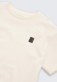 Krämfärgad bomulls-T-shirt med rund halsringning. Har ett litet, mörkbrunt kvadratiskt logomärke på vänster bröstområde.