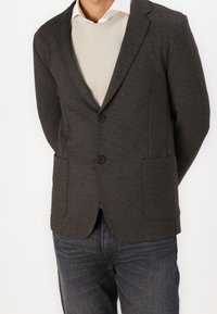 Donkergrijze blazer met een gladde textuur, met notch-lapels, twee voorkantzakken en knoopsluiting, gelaagd over een lichtcrème sweater.