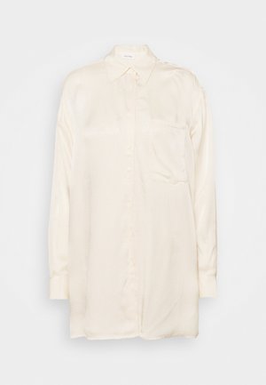 Chemise à manches longues de couleur crème, boutonnée, avec un col pointu et une poche poitrine, présentée sur un fond blanc uni.