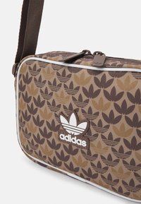 Brązowa torebka na ramię z tekstyliów z nasyconym wzorem liści, białymi akcentami i wytłoczonym logo Adidas z przodu. Zapięcie na zamek.
